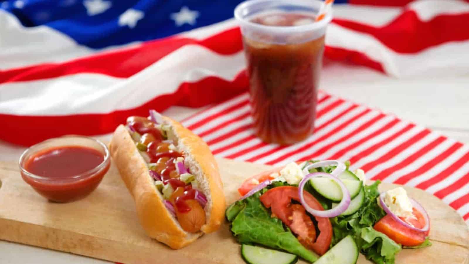 A hot dog with ketchup and onions, a fresh salad, a cup of soda, and a side of ketchup are placed on a wooden board with an American flag backdrop.