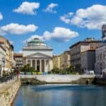 A canal runs through a city lined with pastel buildings, outdoor cafes, and a domed neoclassical church under a blue sky with clouds—an inviting scene perfect for those seeking offbeat travel experiences.