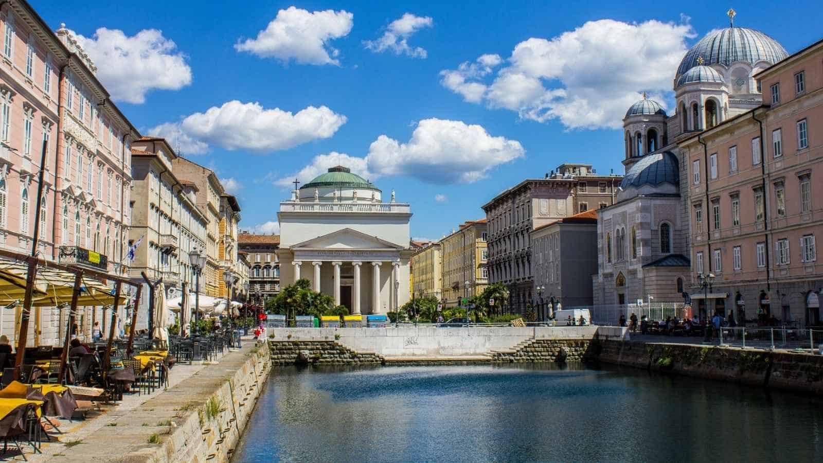 A canal runs through a city lined with pastel buildings, outdoor cafes, and a domed neoclassical church under a blue sky with clouds—an inviting scene perfect for those seeking offbeat travel experiences.