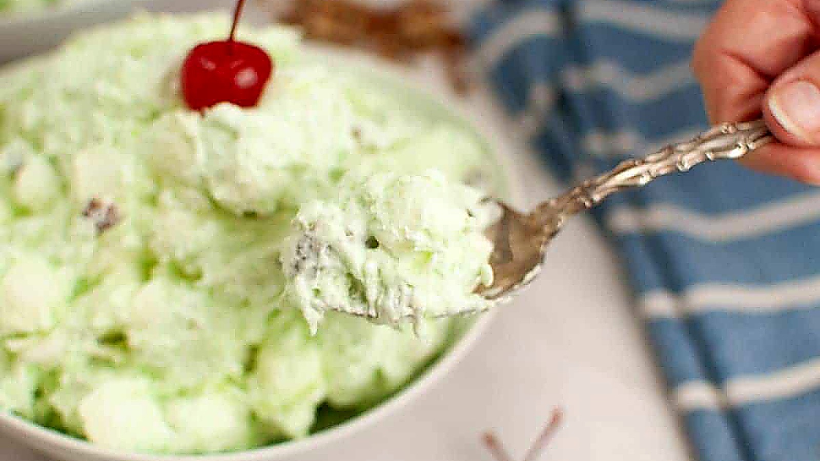 A spoonful of Watergate salad garnished with a cherry, is held over a bowl.