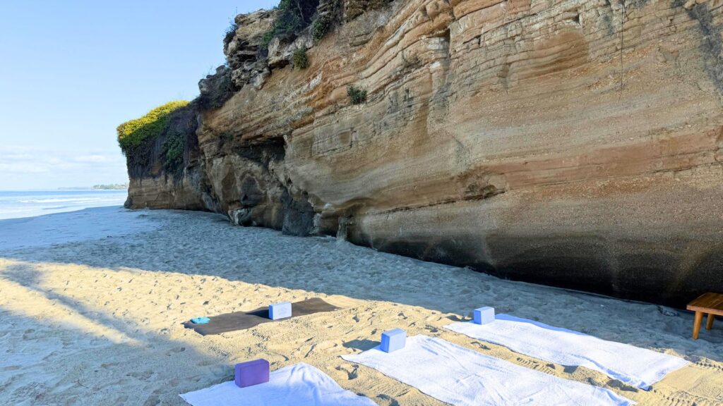 Yoga mats and towels with blocks are set up on sandy beach next to a large rock cliff, with the ocean visible in the background.