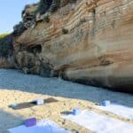 Yoga mats and towels with blocks are set up on sandy beach next to a large rock cliff, with the ocean visible in the background.