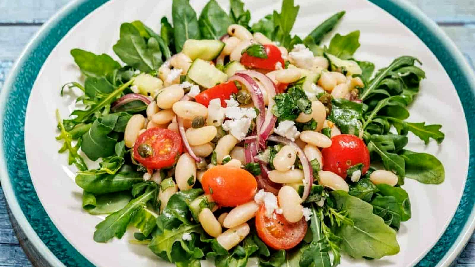 A plate of White Bean Salad with arugula, white beans, cherry tomatoes, red onion, cucumber, and crumbled cheese.