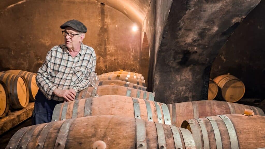 A man in glasses and a cap stands beside wooden barrels in a dimly lit wine cellar with stone walls and an arched ceiling, evoking the ambiance of a southern France barge cruise.
