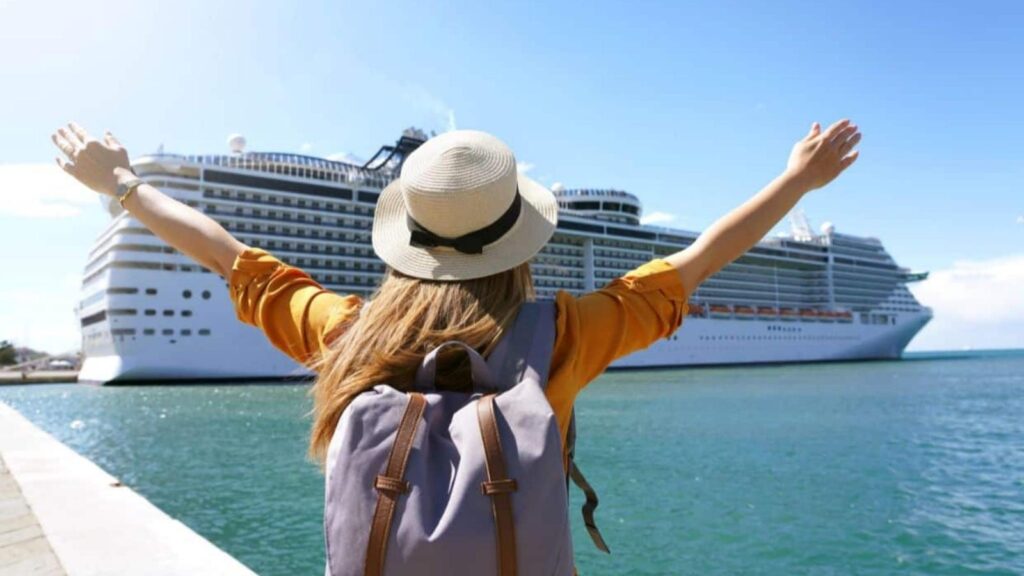 A person with a backpack and hat raises their arms, facing a large white cruise ship docked at the port—ready for an unforgettable Fourth of July travel adventure.