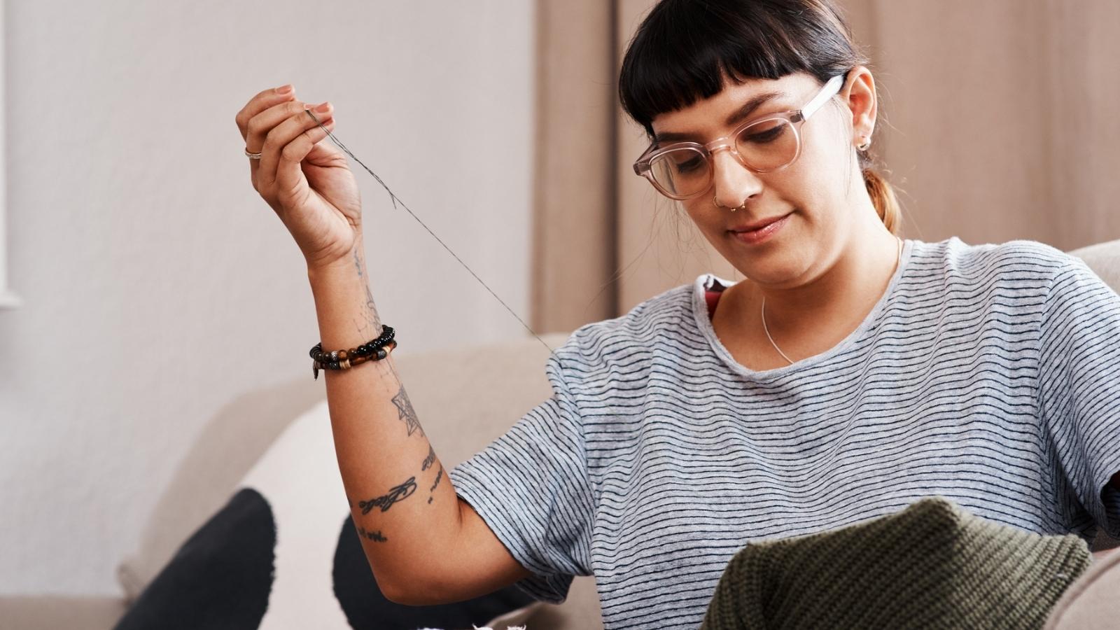 Person with glasses threading a needle while holding dark green fabric, embodying simple living as they sit on a sofa in a well-lit room.