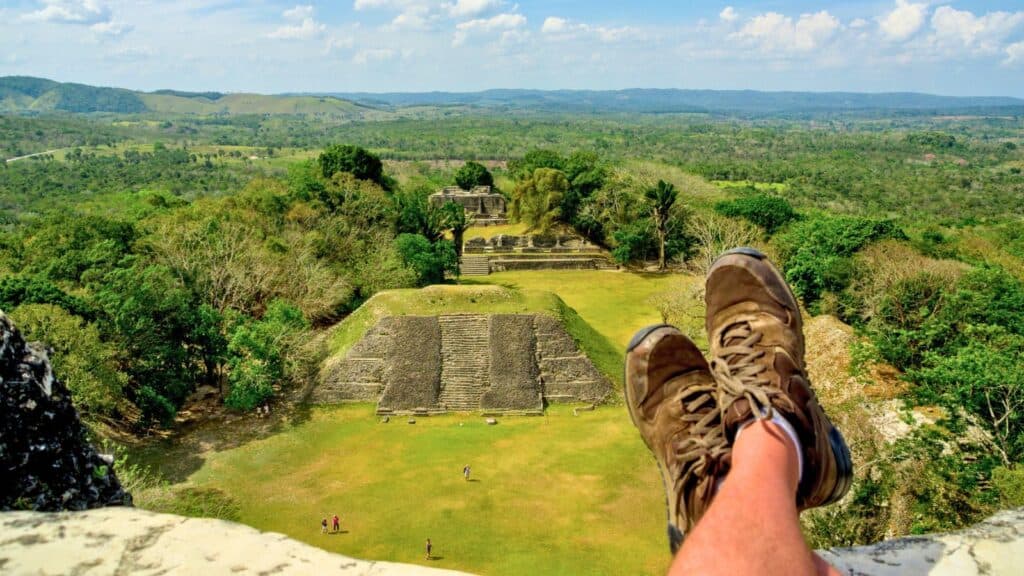 A person with their legs outstretched sits above ancient stone ruins, perhaps pausing during a Belize reef and ruins trip, surrounded by green landscape and distant hills under a blue sky with scattered clouds.