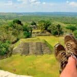 A person with their legs outstretched sits above ancient stone ruins, perhaps pausing during a Belize reef and ruins trip, surrounded by green landscape and distant hills under a blue sky with scattered clouds.