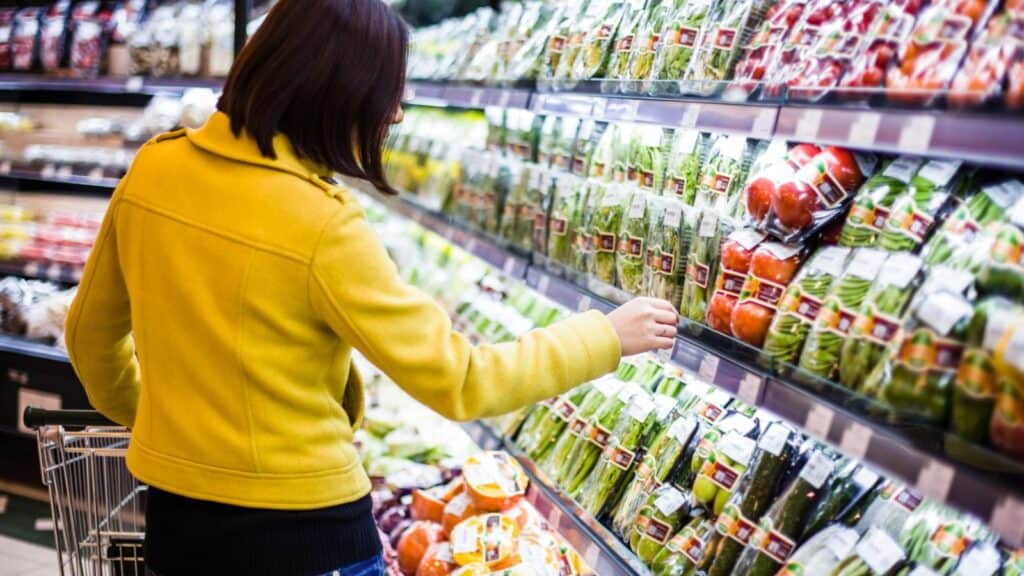 Person wearing a yellow jacket selects packaged produce from a grocery store shelf, with various fresh vegetables and fruits displayed in plastic containers.