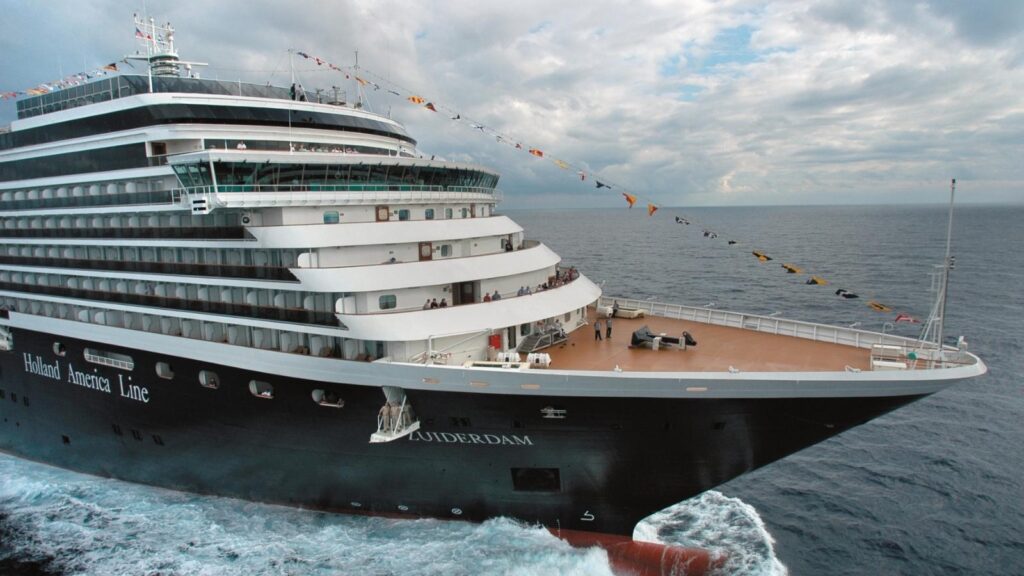 A large Holland America Line cruise ship travels through the ocean under a cloudy sky, with colorful signal flags strung above its decks.