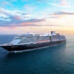 A large cruise ship sails on calm ocean waters at sunset under a partly cloudy sky.