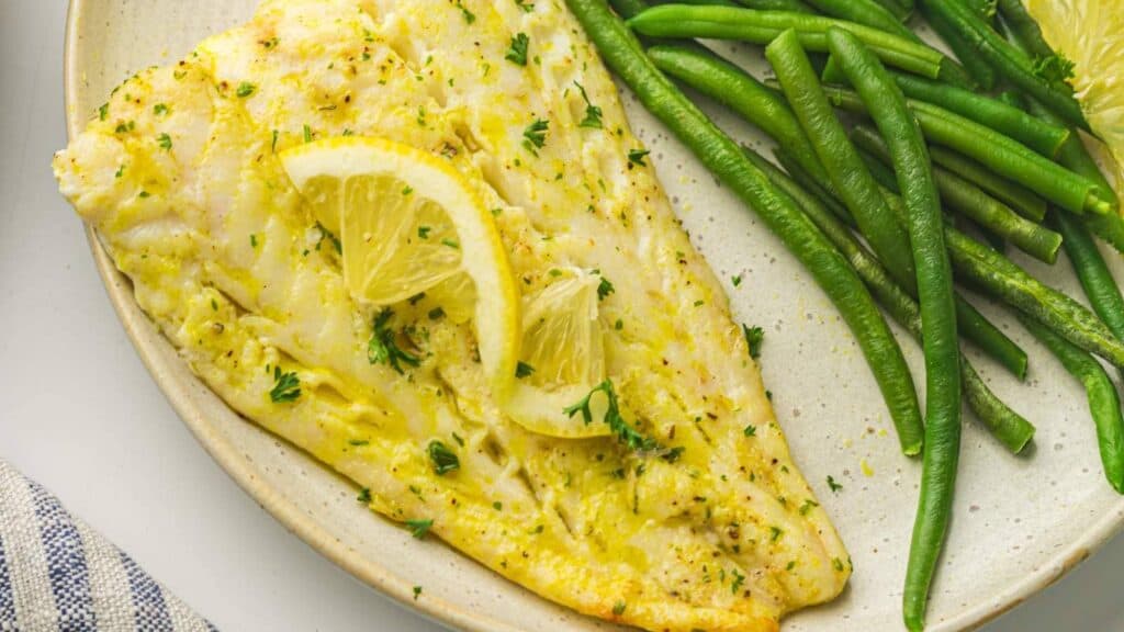A plate with a baked white fish fillet garnished with lemon slices and herbs, served alongside cooked green beans.