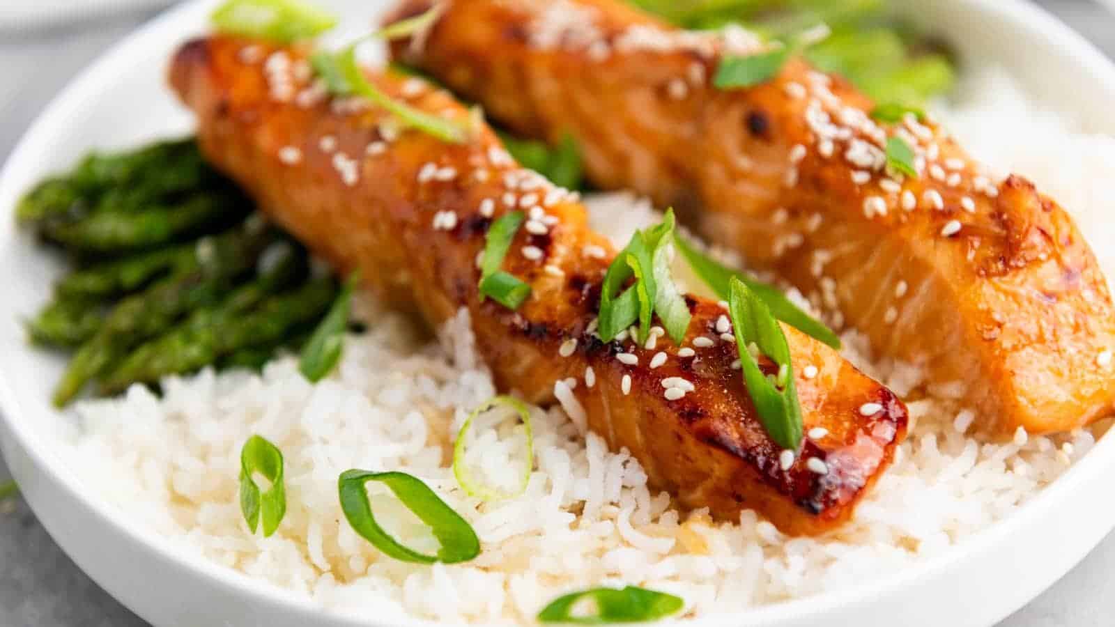 Two glazed salmon fillets topped with sesame seeds and sliced green onions served over white rice with a side of asparagus.