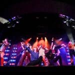 A group of performers in colorful, retro-style stage costumes dance and sing under bright spotlights on a dark stage.