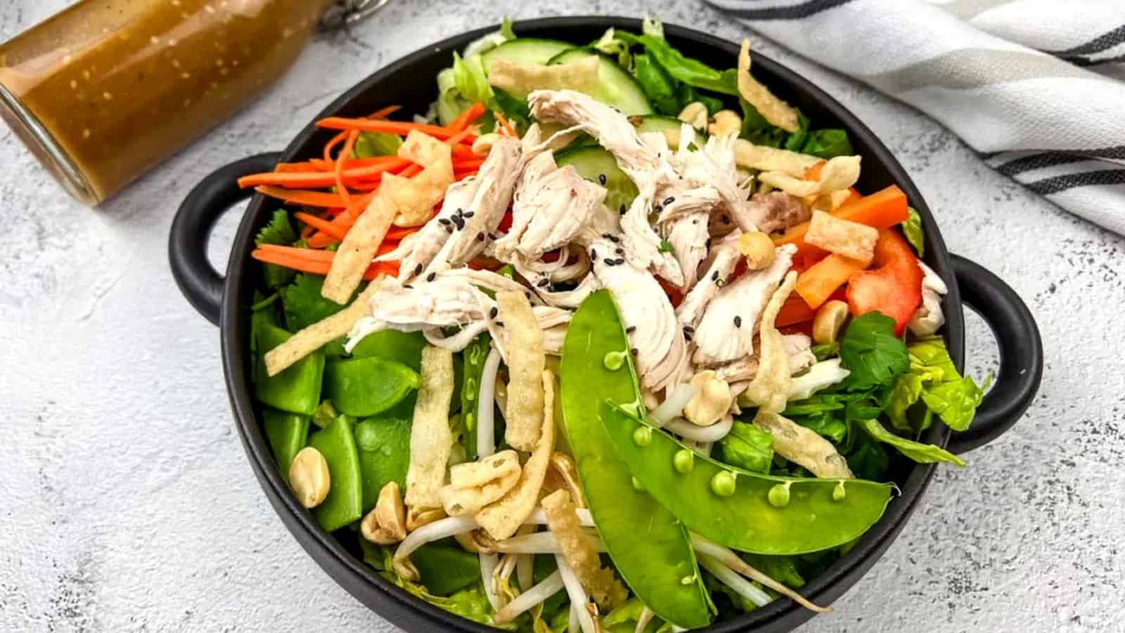 A bowl of Chinese chicken salad with shredded chicken, snap peas, carrots, and peppers, accompanied by a bottle of dressing on the side.