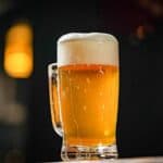 A clear glass mug filled with golden beer and a thick foamy head, set against a dark, blurred background with warm lighting.
