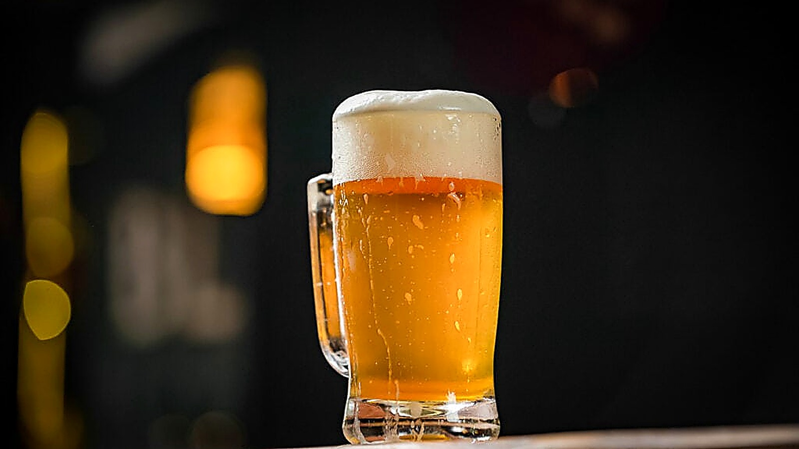 A clear glass mug filled with golden beer and a thick foamy head, set against a dark, blurred background with warm lighting.