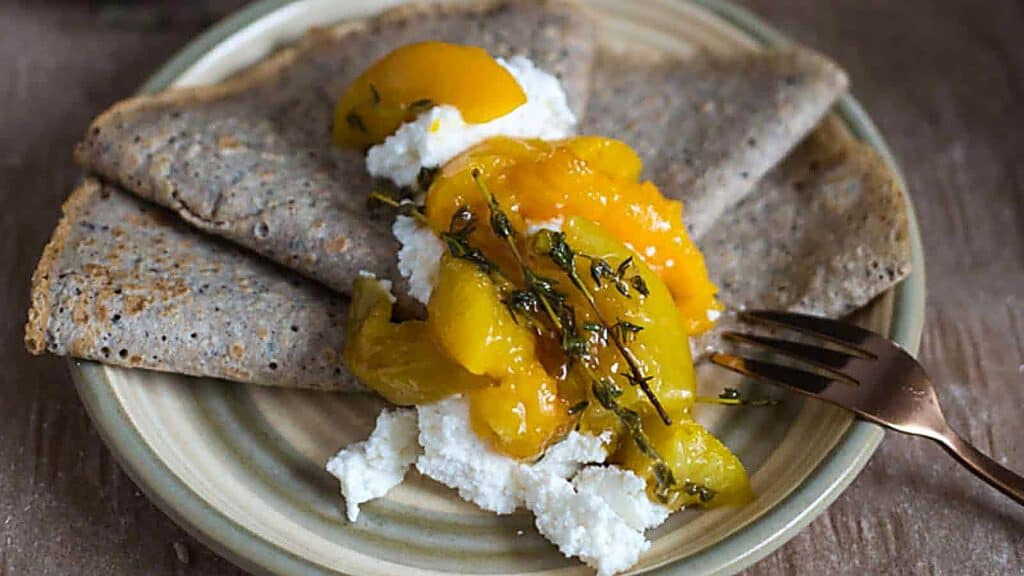Two folded crepes on a plate topped with ricotta cheese, sliced peaches, and fresh herbs, next to a gold fork.