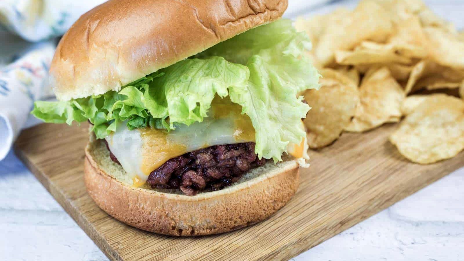 A smoked burger with lettuce and a slice of cheddar cheese on a bun, served with potato chips on a wooden board.