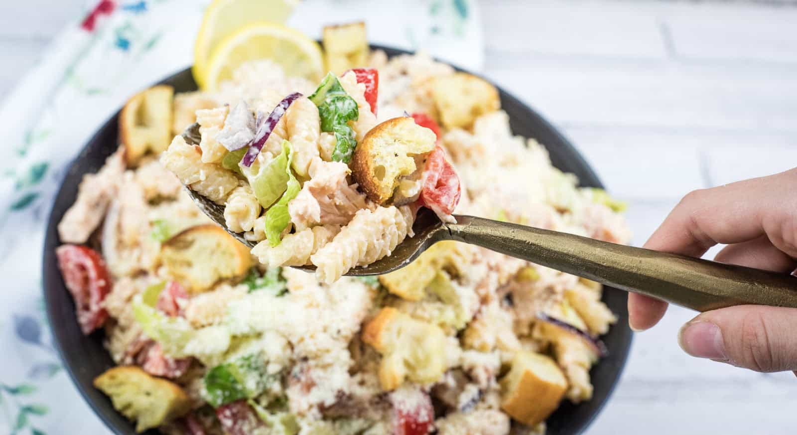 A hand holds a fork with pasta salad, croutons, lettuce, and tomatoes above a bowl of the same salad on a white surface.