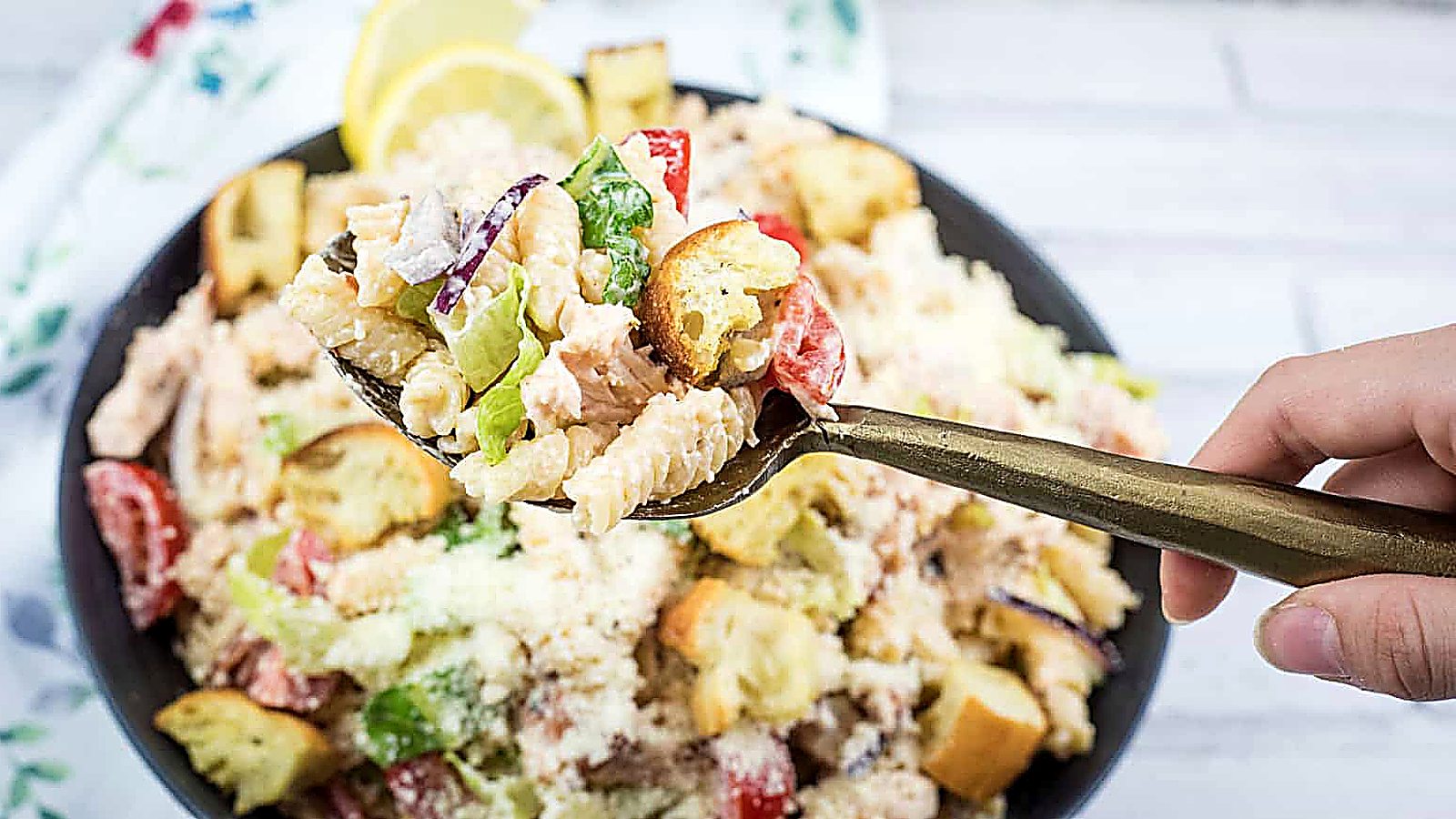 A hand holds a fork with pasta salad, croutons, lettuce, and tomatoes above a bowl of the same salad on a white surface.