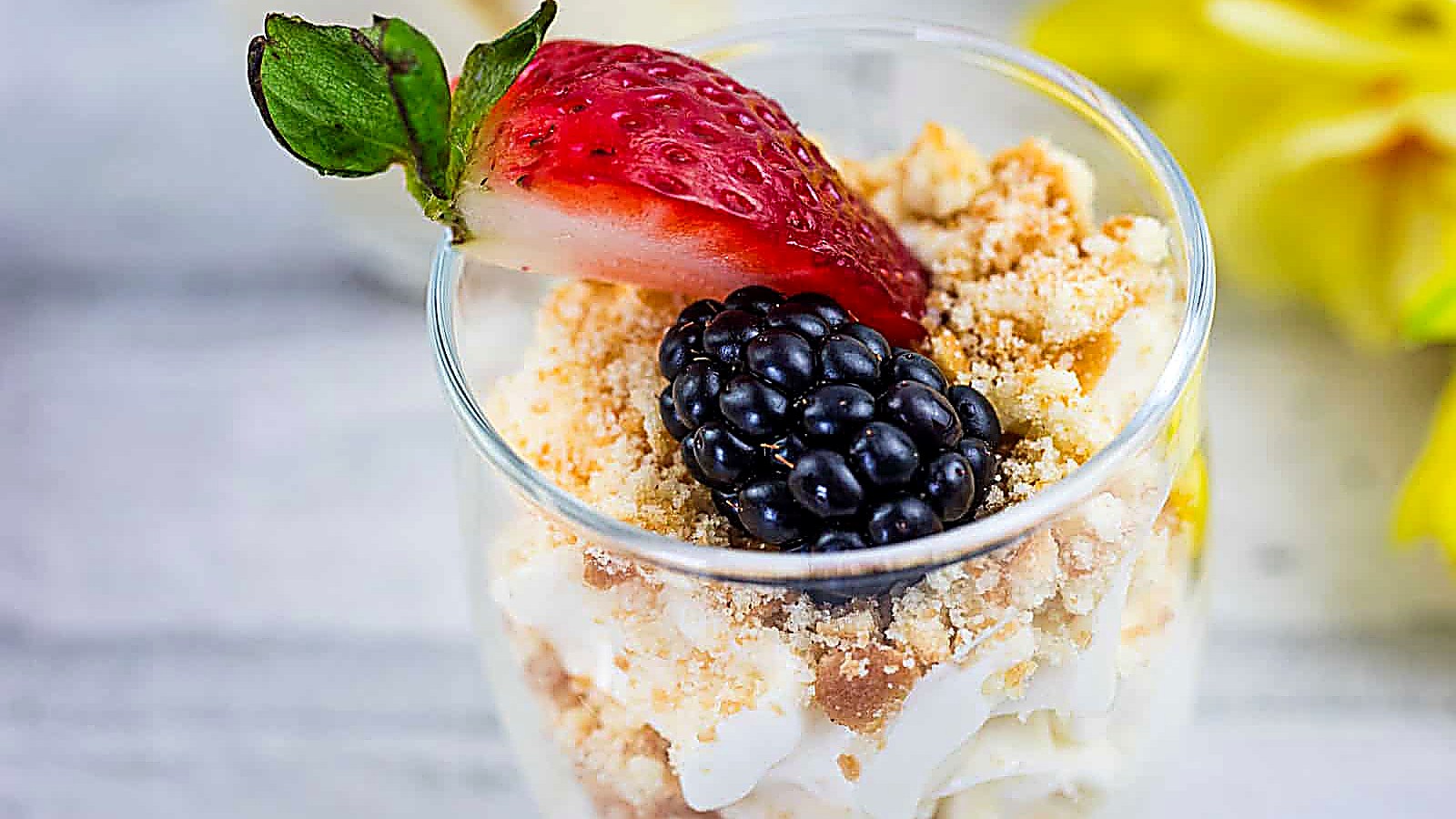 A dessert cup filled with crumbled cookies, cream, topped with a sliced strawberry and a blackberry, set on a light surface with yellow flowers in the background.