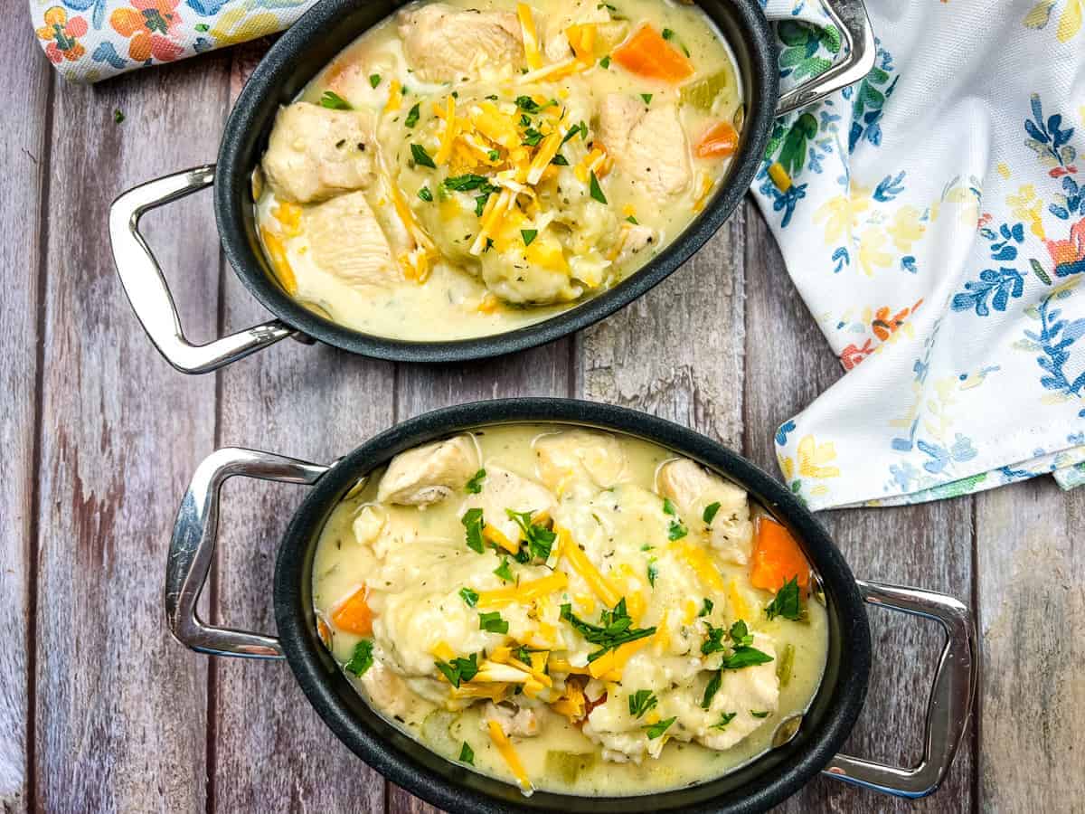 Two black oval dishes filled with creamy chicken stew, vegetables, and topped with shredded cheese and herbs, placed on a wooden surface near a floral napkin.