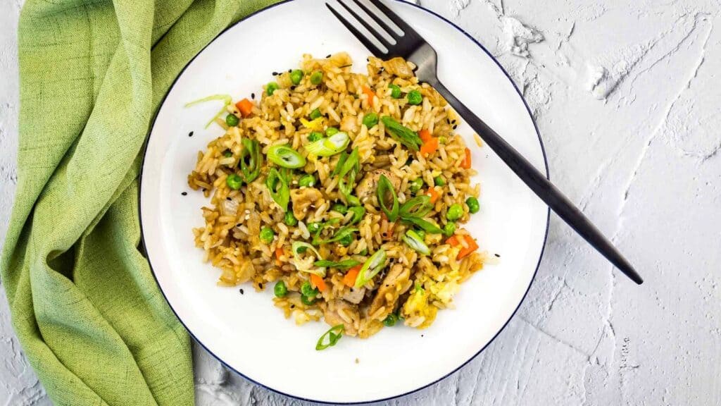 A plate of fried rice with vegetables, garnished with sliced green onions, sits on a white surface with a black fork and a green cloth napkin nearby.