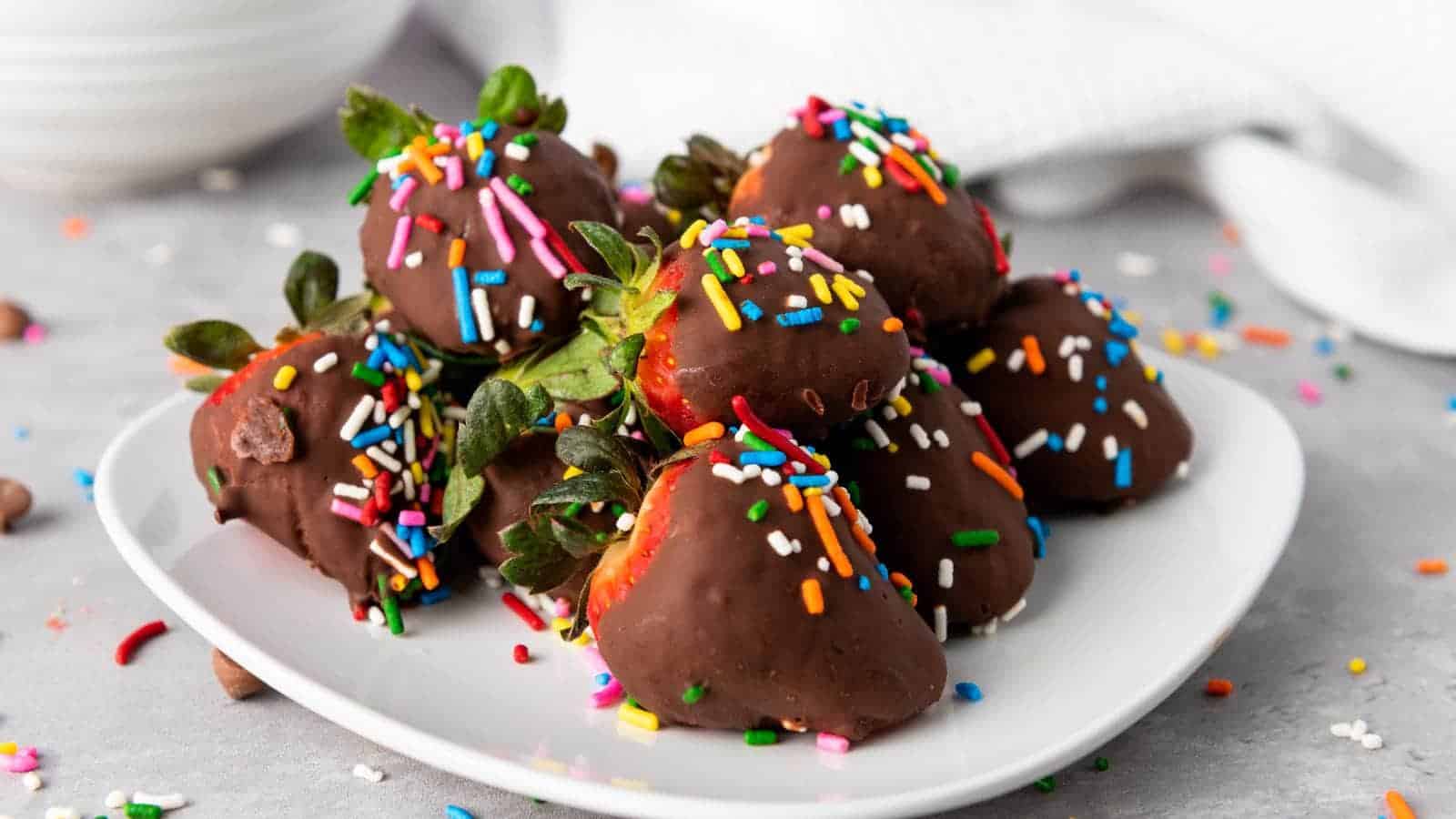 A white plate holds several chocolate-covered strawberries topped with colorful sprinkles, with extra sprinkles scattered on the gray surface nearby.