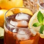 A glass of iced tea with ice cubes, garnished with a lemon slice and mint sprig, with whole lemons and a green cloth in the background.