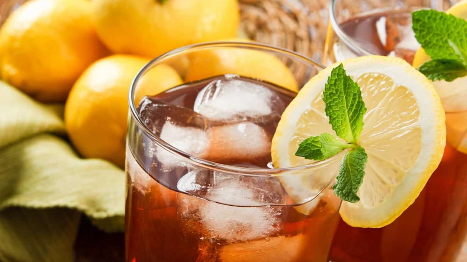 A glass of iced tea with ice cubes, garnished with a lemon slice and mint sprig, with whole lemons and a green cloth in the background.