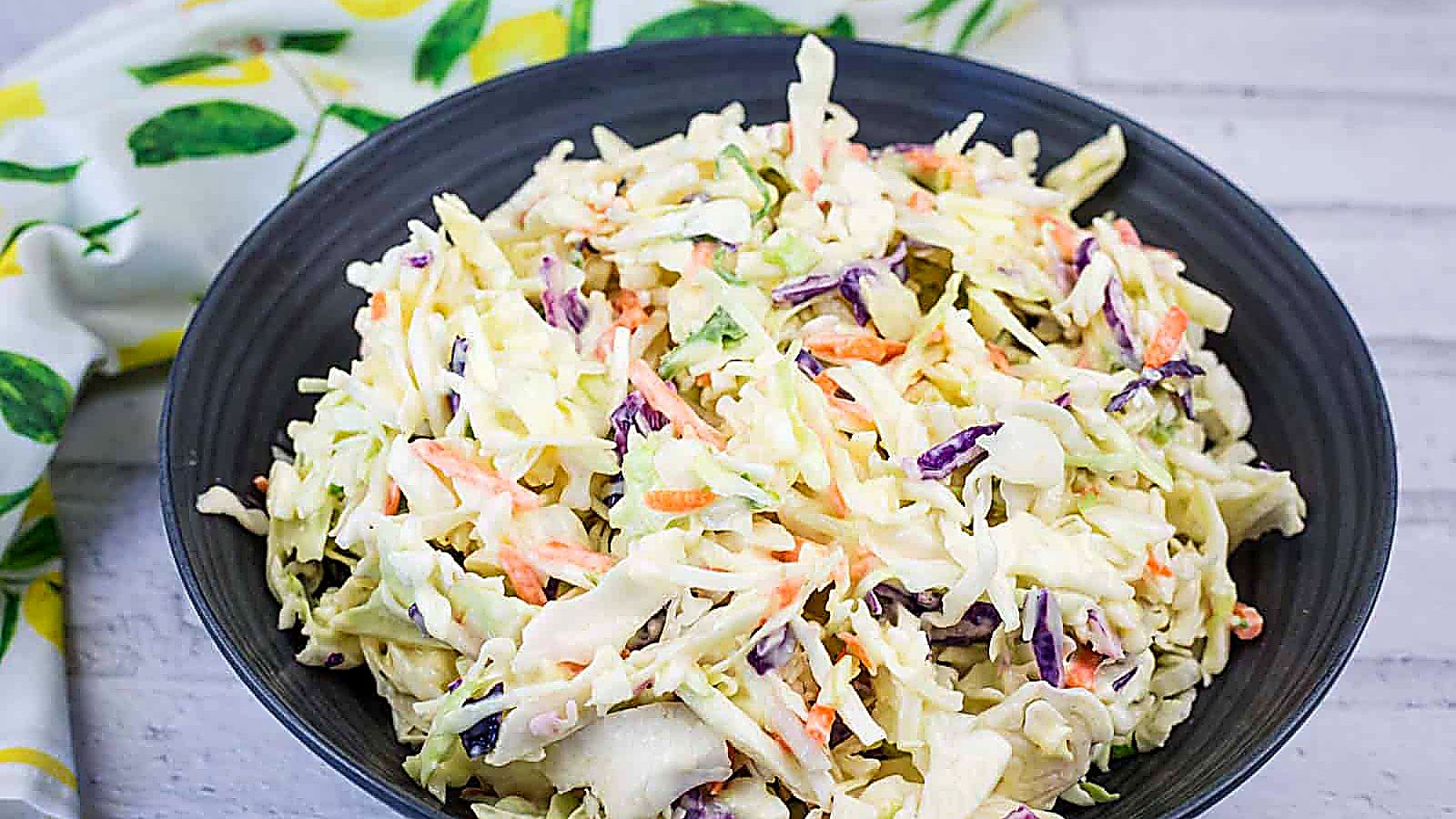 A black bowl filled with creamy coleslaw made of shredded cabbage, carrots, and purple cabbage, placed on a light-colored surface with a patterned cloth nearby.