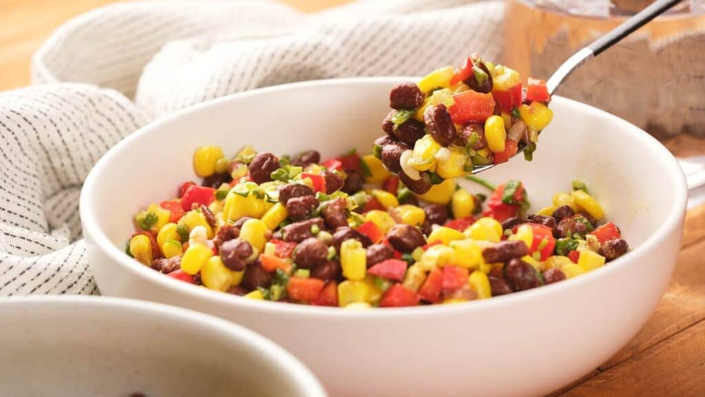 A white bowl filled with a colorful salad of black beans, corn, diced red bell pepper, and chopped herbs, with a spoon lifting a portion.