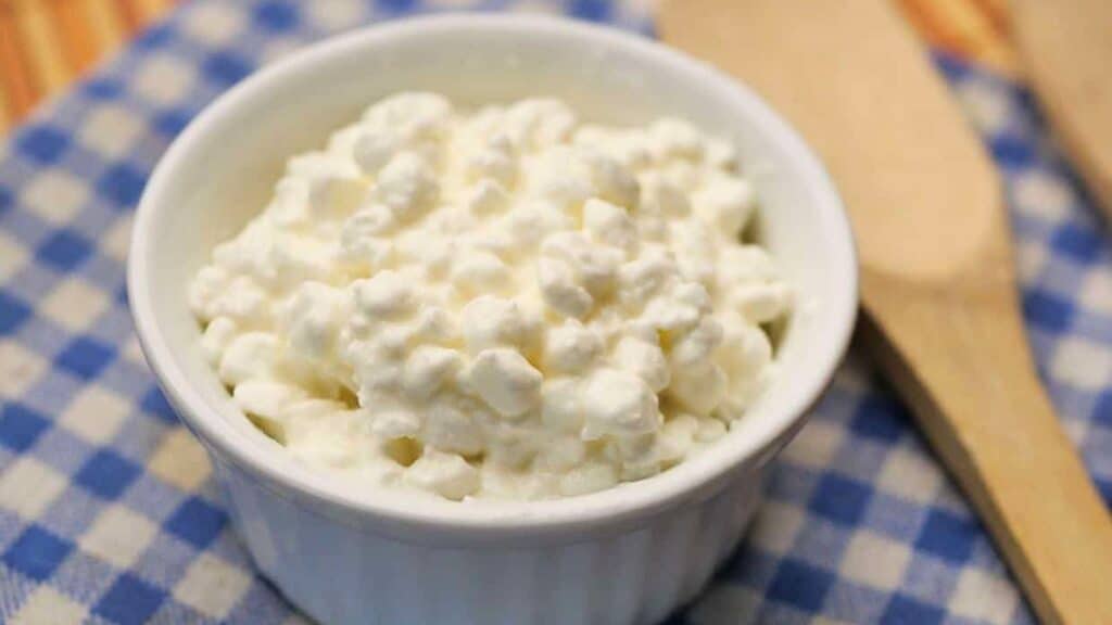 A small white bowl filled with cottage cheese sits on a blue and white checkered cloth next to a wooden utensil.