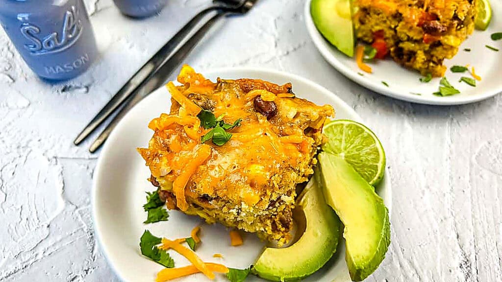 A square slice of cheesy casserole on a white plate, garnished with avocado slices, lime wedge, and chopped herbs, with utensils and salt shaker nearby.