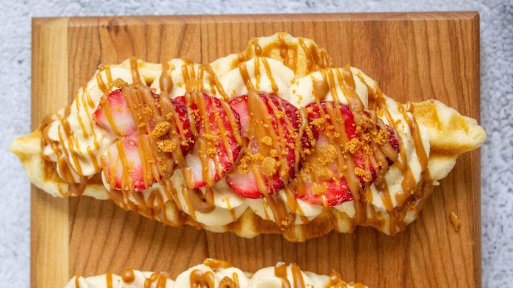 A waffle topped with cream, sliced strawberries, crumbled biscuit, and caramel drizzle, served on a wooden board&mdash;a delightful addition to your collection of breakfast treats.