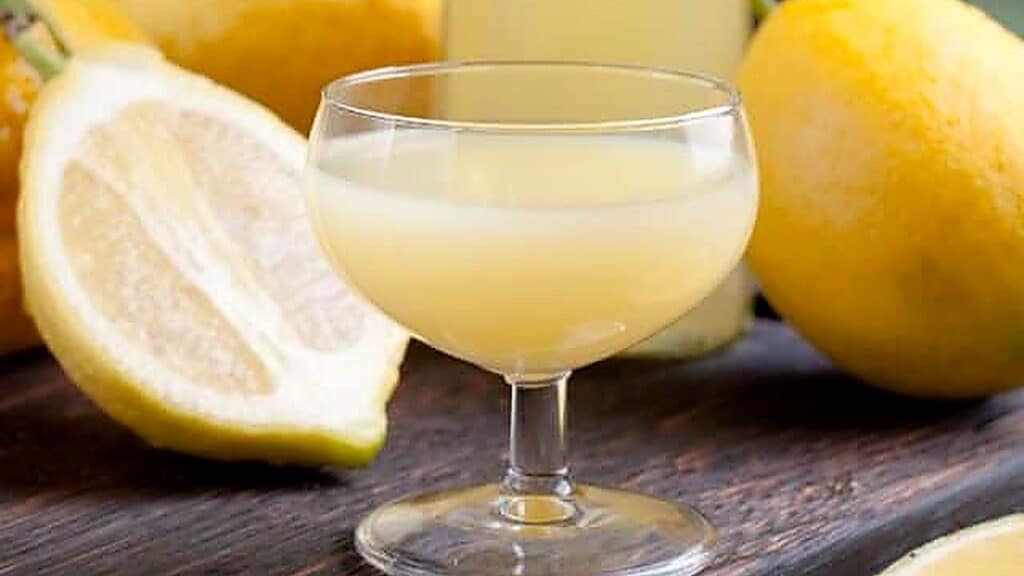 A coupe glass filled with pale yellow lemon juice sits on a wooden surface, surrounded by whole and sliced lemons.