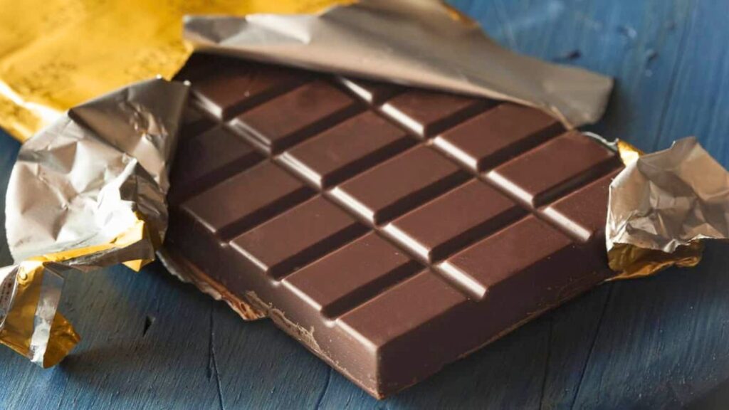 A partially unwrapped dark chocolate bar lies on a blue wooden surface, with gold and silver foil pulled back to reveal the segmented chocolate.