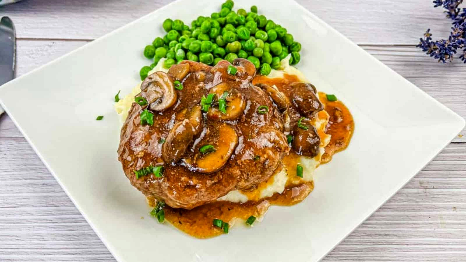 A white plate with Salisbury steak topped with mushroom gravy on mashed potatoes, accompanied by a side of green peas.