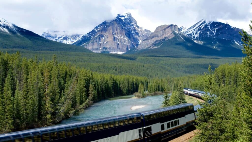 A passenger train travels along a river through a forested valley with snow-capped mountains in the background under a cloudy sky.