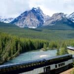 A passenger train travels along a river through a forested valley with snow-capped mountains in the background under a cloudy sky.