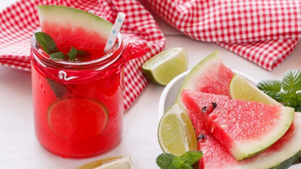 Celebrate National Watermelon Month with a refreshing glass of red watermelon drink, garnished with lime slices and mint, beside a plate of juicy watermelon wedges, lime wedges, and a red checkered cloth.