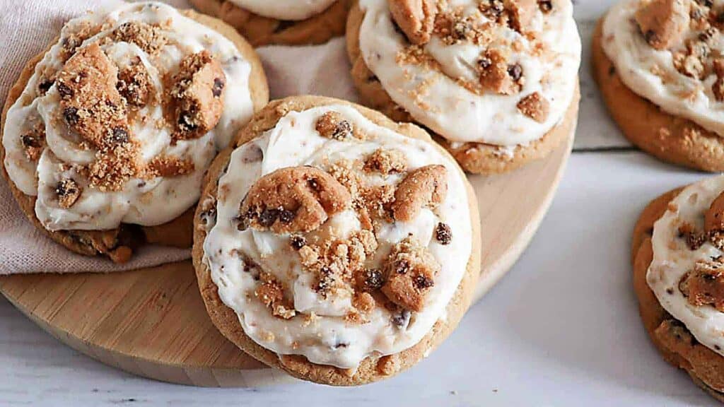 Cookies topped with a creamy frosting and mini chocolate chip cookies, arranged on a wooden board and a light fabric.