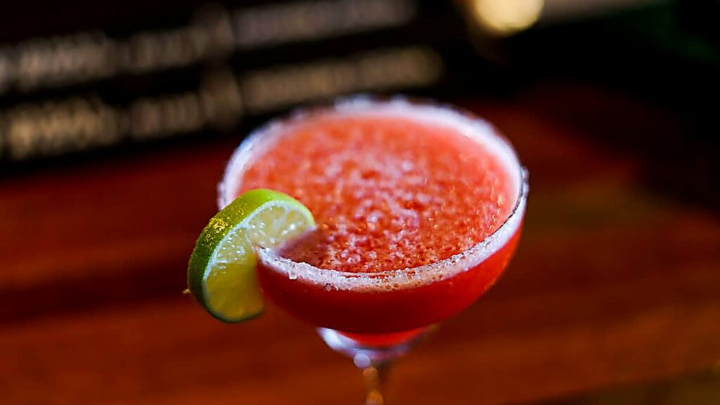 A red frozen cocktail in a coupe glass, garnished with a lime wedge on the rim, sits on a wooden surface—showcasing one of the vibrant margarita flavors perfect for summer celebrations.