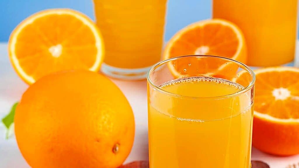 A glass of orange juice is in the foreground, surrounded by whole and halved oranges and additional glasses of juice in the background.