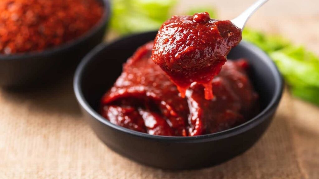 A close-up of a spoonful of red chili paste being lifted from a black bowl, with another bowl of spices and green garnish in the background.