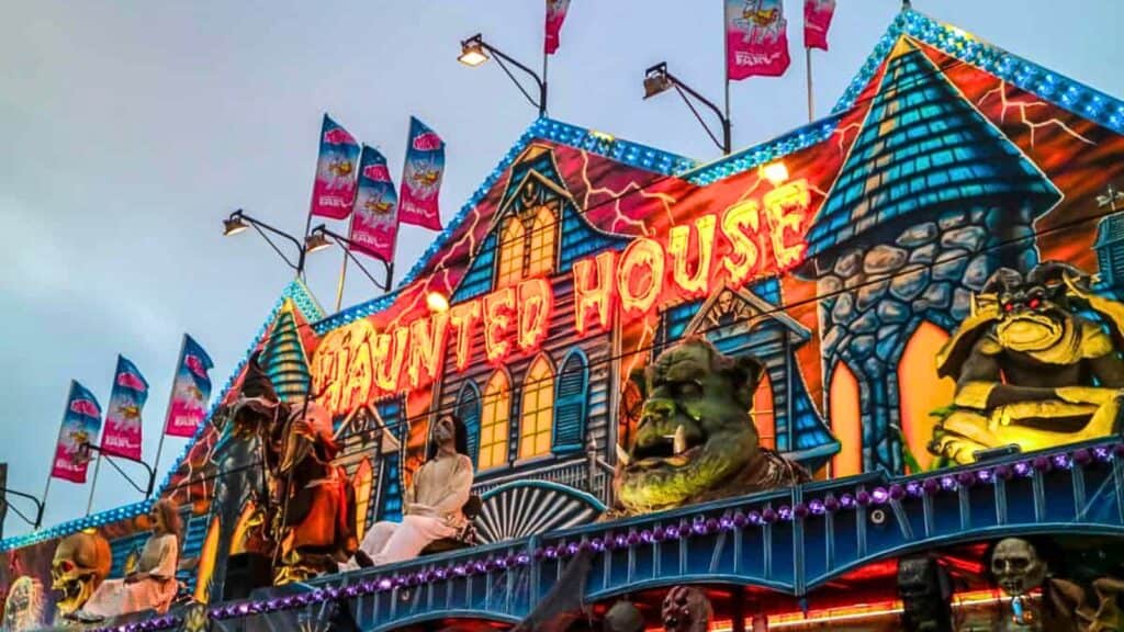 Colorful haunted house attraction at a fair, featuring bright lights, Halloween decorations, monstrous figures, and several flags on the roof against a cloudy sky.
