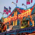 Colorful haunted house attraction at a fair, featuring bright lights, Halloween decorations, monstrous figures, and several flags on the roof against a cloudy sky.