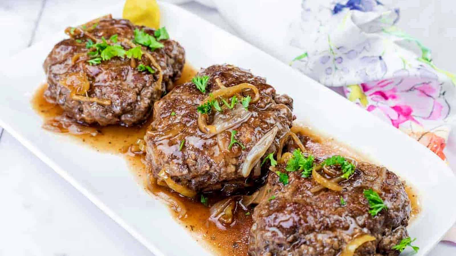 Three Salisbury steak patties topped with onions and brown gravy, garnished with parsley, served on a white rectangular plate.