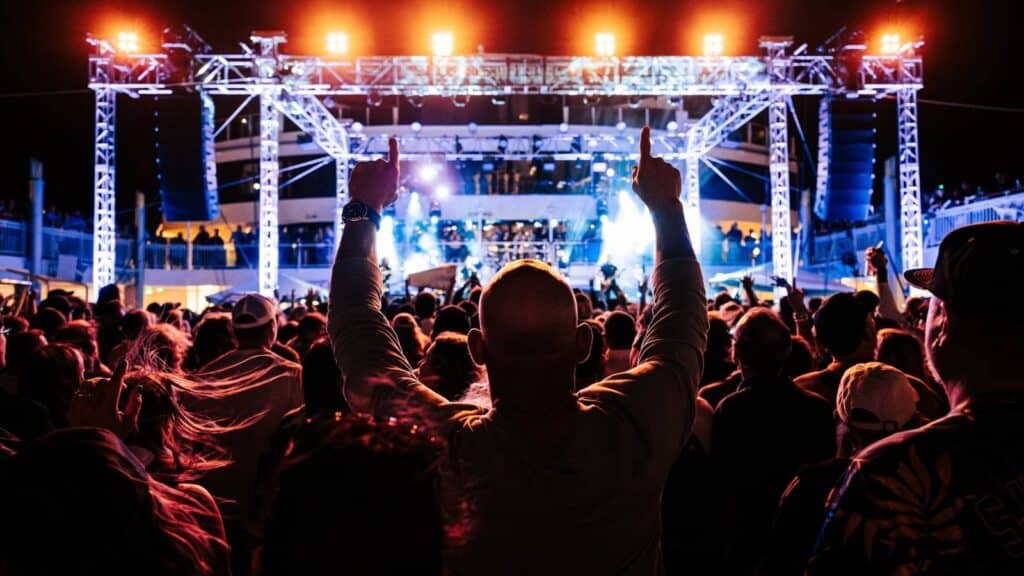 A crowd watches an outdoor concert at night, with bright stage lights illuminating performers and a person in the foreground raising their hands.